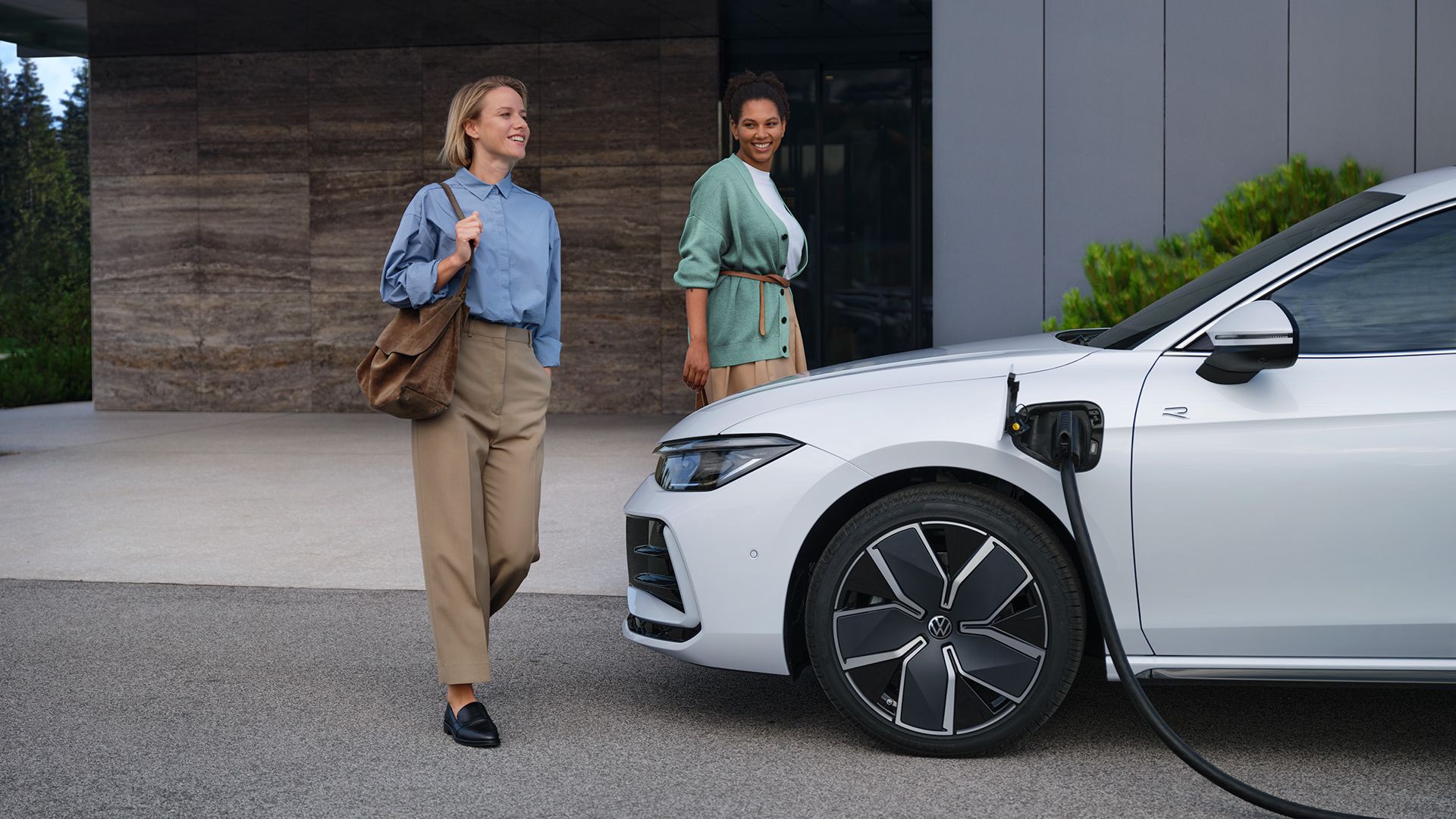 VW Passat in weiß seitlich vor einem Bürogebäude mit eingestecktem Ladekabel, zwei Frauen gehen auf den Wagen zu.