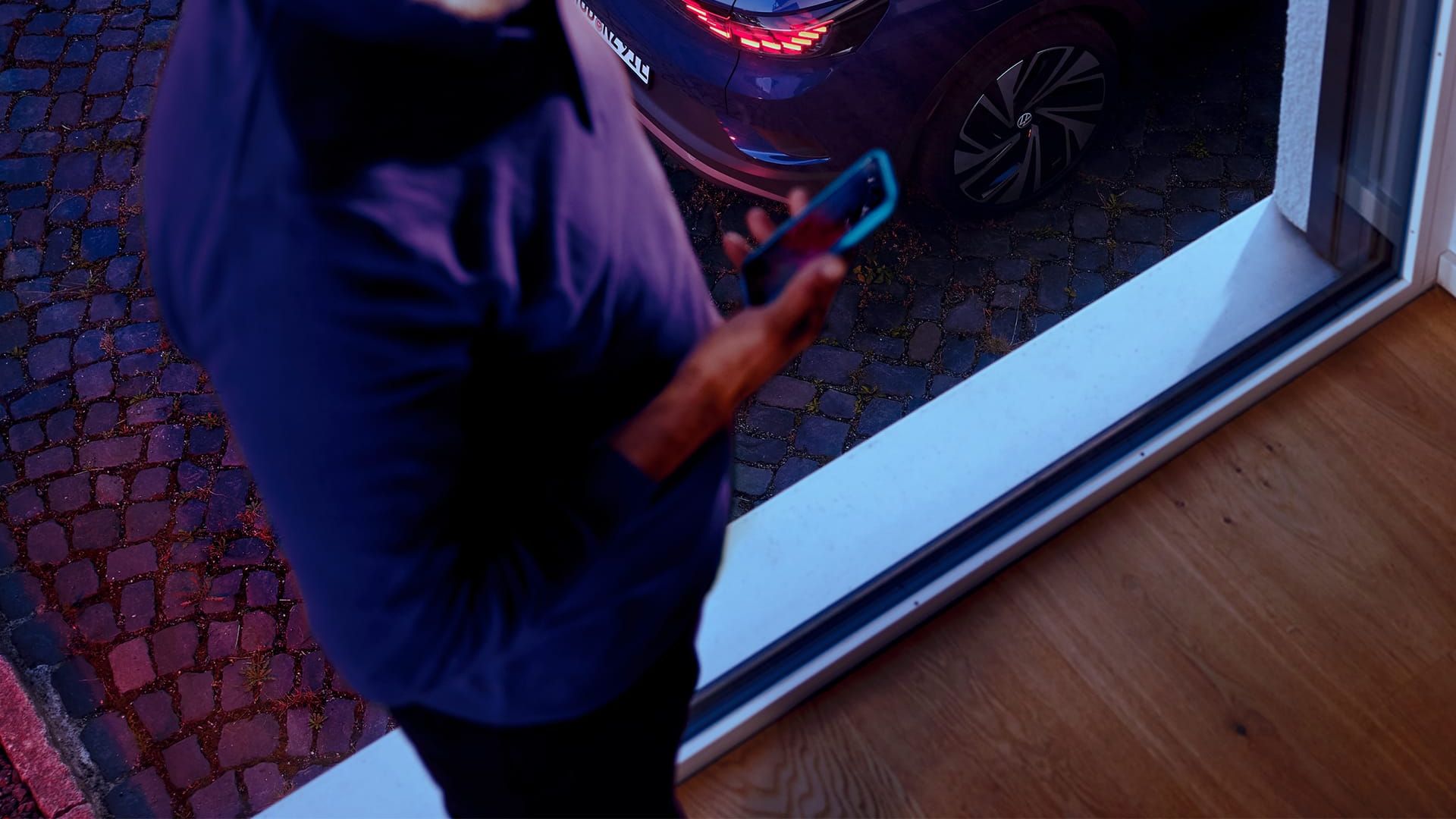 Mann mit Smartphone in der Hand steht am Fenster, im Hintergrund steht ein blauer VW ID.4