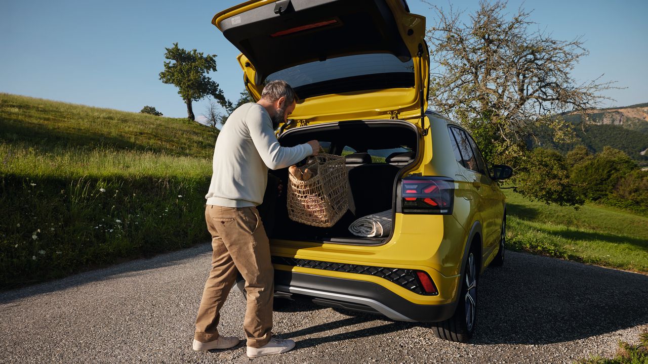 Ein Mann stellt einen großen Picknickkorb in den Kofferraum eines gelben VW T-Cross, der auf einer Straße in hügeliger Landschaft steht.