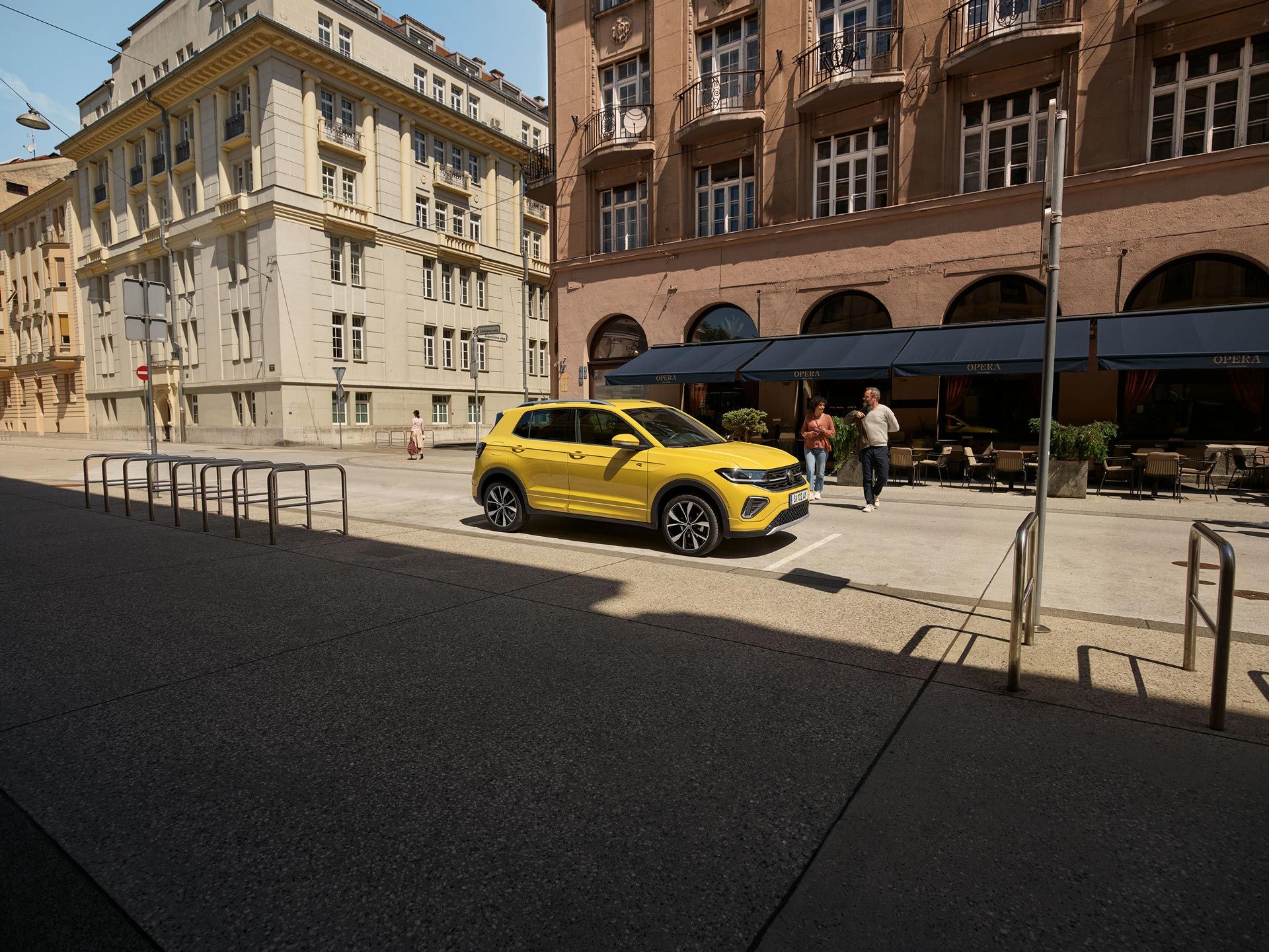 VW T-Cross in gelb parkt seitlich vor einem urbanen Straßencafé, ein Päarchen geht auf den Wagen zu.
