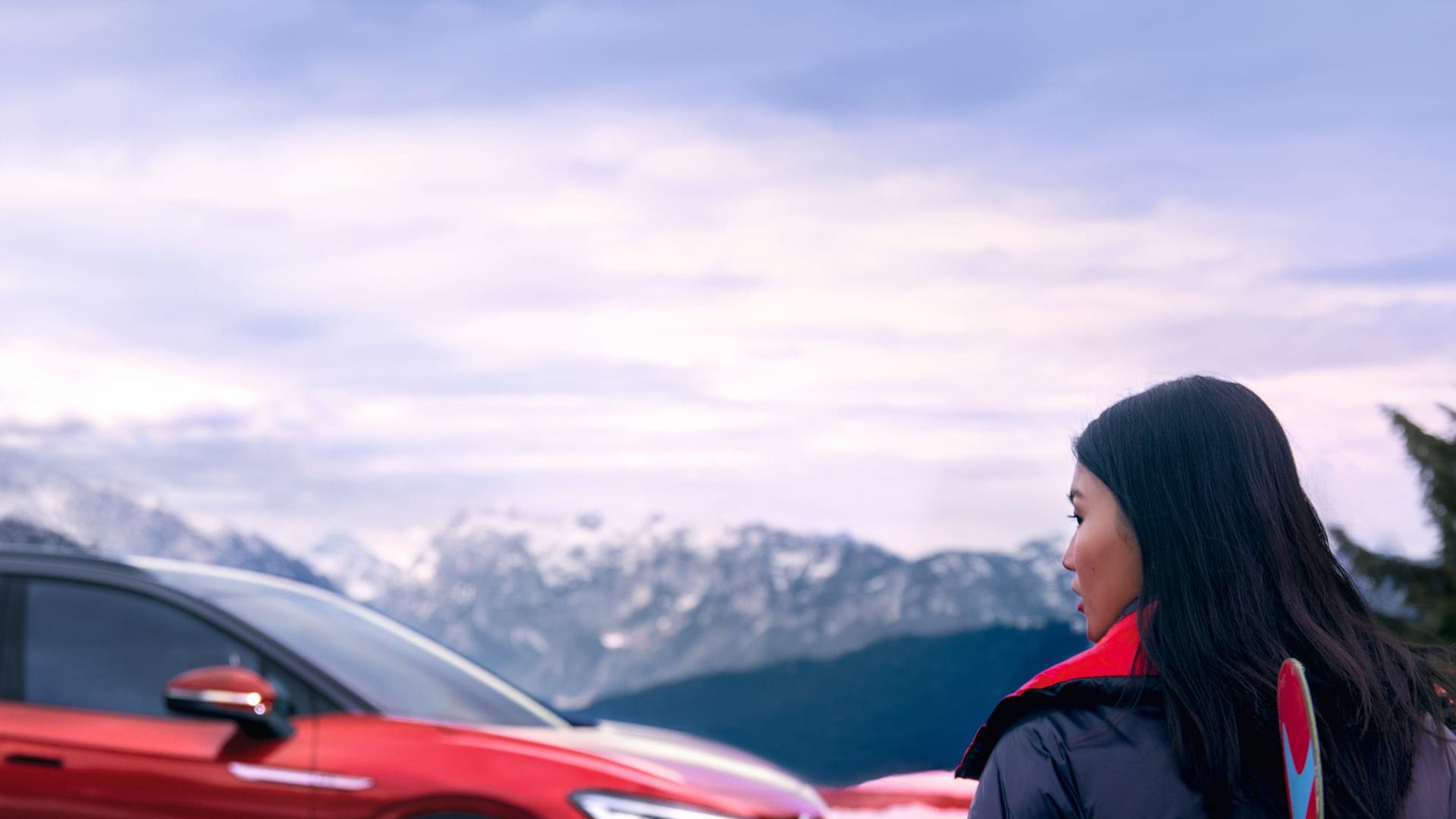 Seitenansicht eines roten VW ID.4 GTX vor Bergkulisse, im Vordergrund blickt eine Frau zum Auto