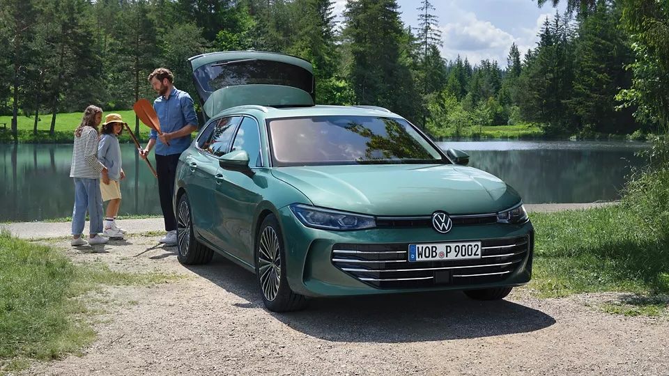 Der neue VW Passat in blaugrün parkt an einem Badesee mit geöffnetem Kofferraum. Ein Mann verteilt Freitzeitspielzeug an zwei Kinder.