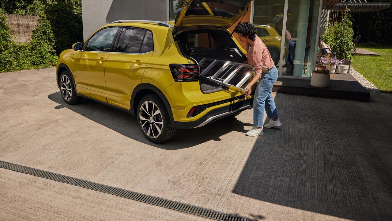 Eine junge Frau schiebt eine Leiter in den Kofferraum eines gelben VW T-Cross.
