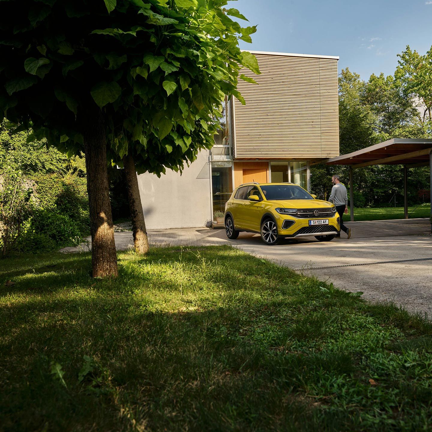 Ein Mann macht sich bereit, in einen gelben VW T-Cross einzusteigen, der in Frontansicht auf einer Garteneinfahrt eines Hauses geparkt ist.