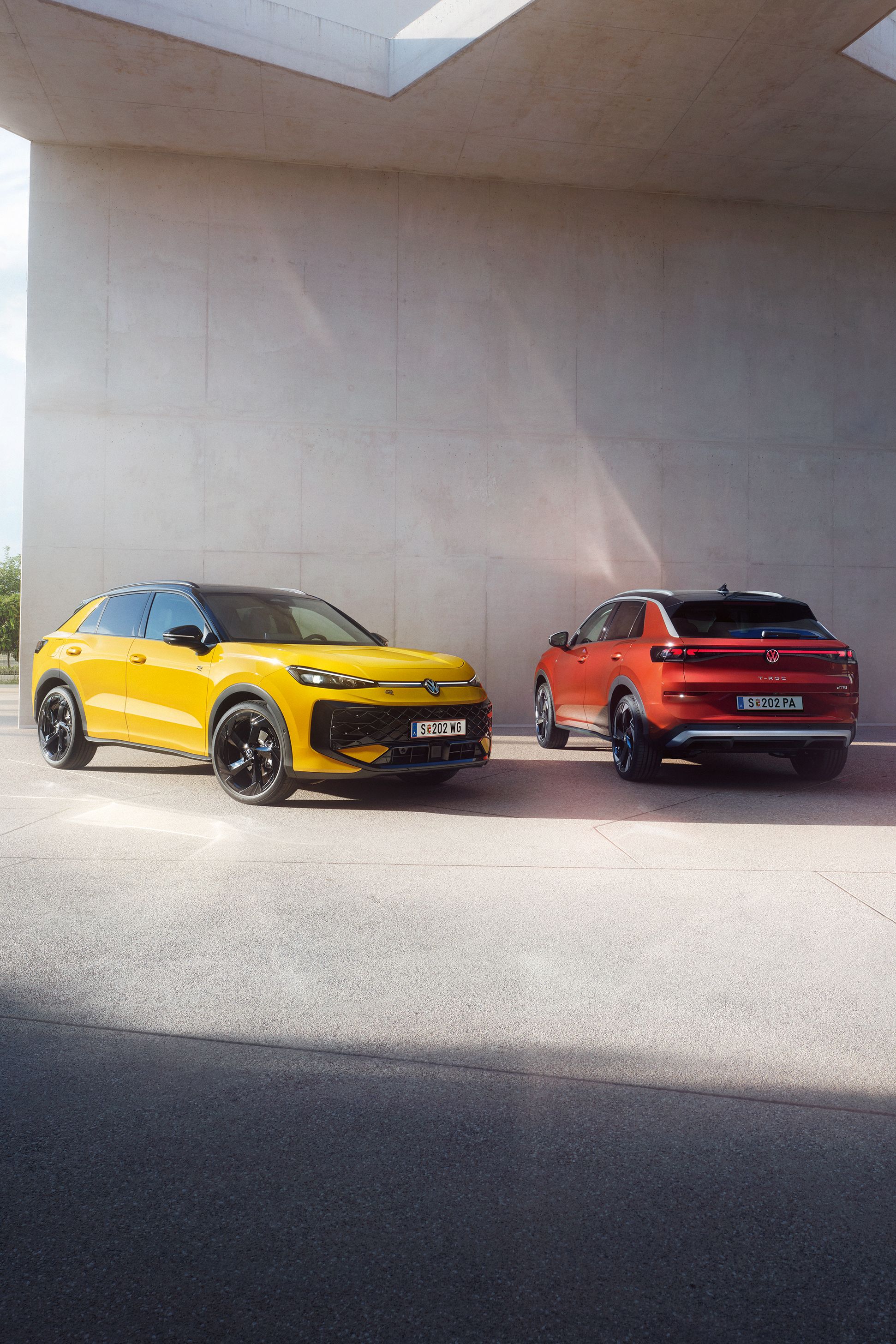 Ein gelber VW T-Roc R-Line und ein roter VW T-Roc Style stehen nebeneinander vor einem modernen Gebäude.