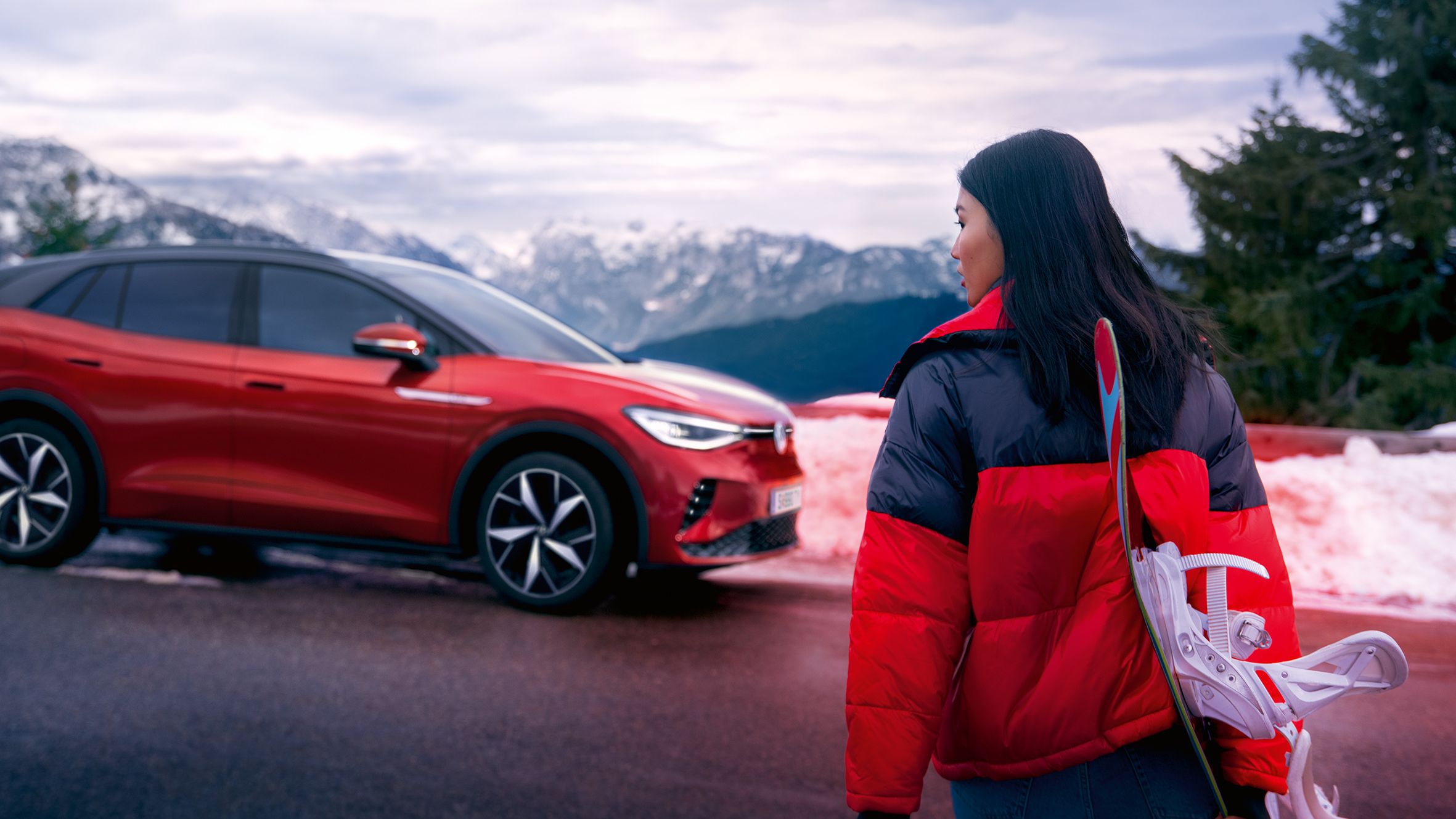 VW ID.4 GTX in rot auf Straße in Berglandschaft mit Schnee. Frau in roter-schwarzer Jacke überquert mit Skiern die Straße und guckt nach links.