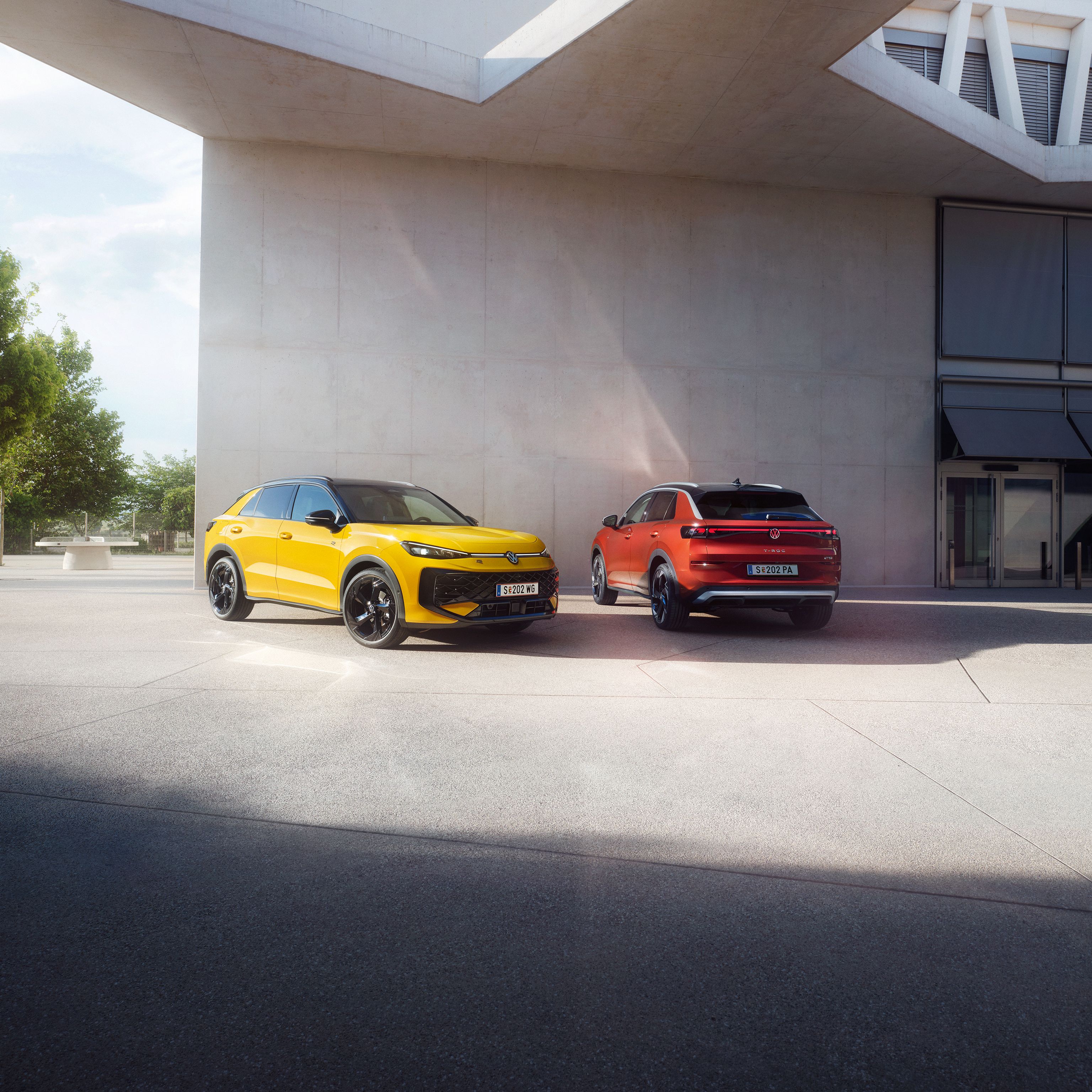 Ein gelber VW T-Roc R-Line und ein roter VW T-Roc Style stehen nebeneinander vor einem modernen Gebäude.