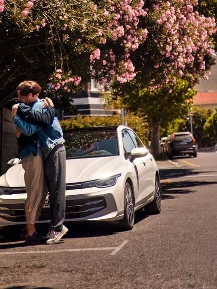 Weißer VW Golf wird an einer Ladestation geladen. Zwei Männer haben sich neben dem Auto in den Armen.
