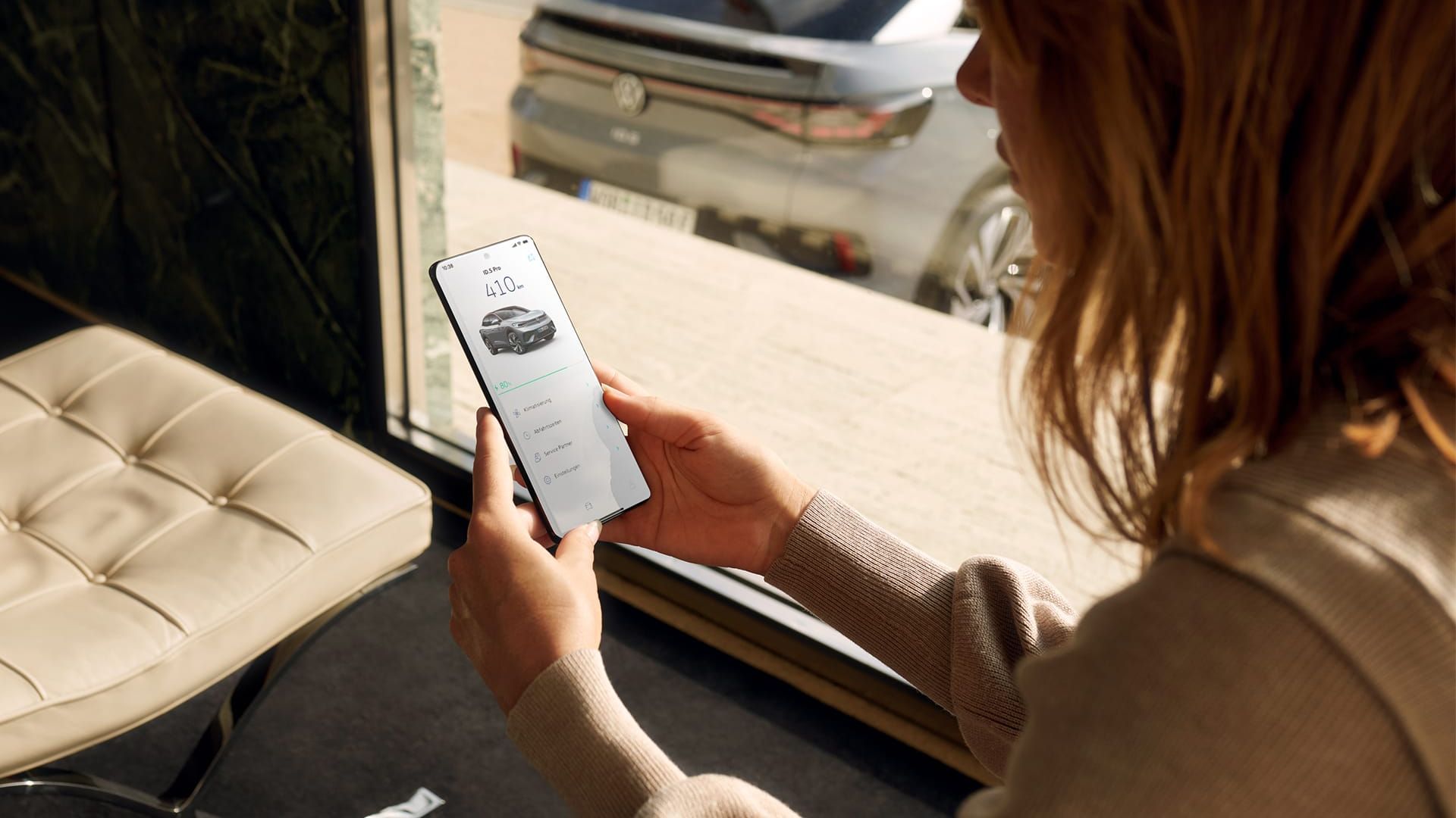 Frau sitzt im Raum und schaut aufs Smartphone, durchs Fenster ist das Heck eines grauen VW ID.5 sichtbar