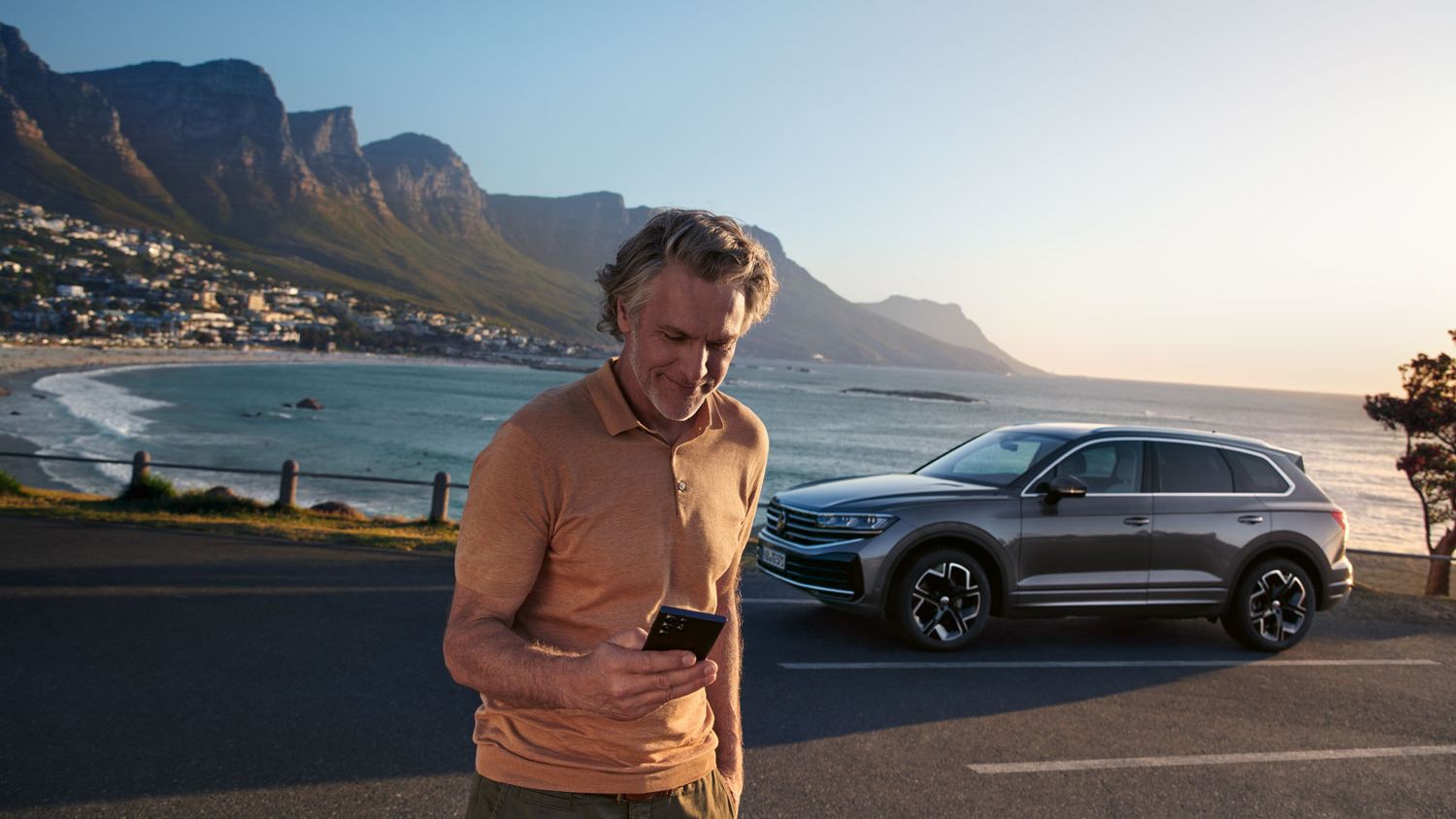 Ein Mann schaut auf sein Smartphone, im Hintergrund steht der Touareg vor einem See.