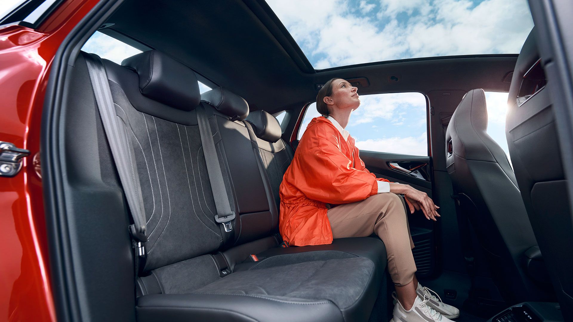 Eine junge Frau sitzt auf der Rückbank eines roten VW ID.5 GTX und schaut durch das Panoramaglasdach in den blauen Himmel.