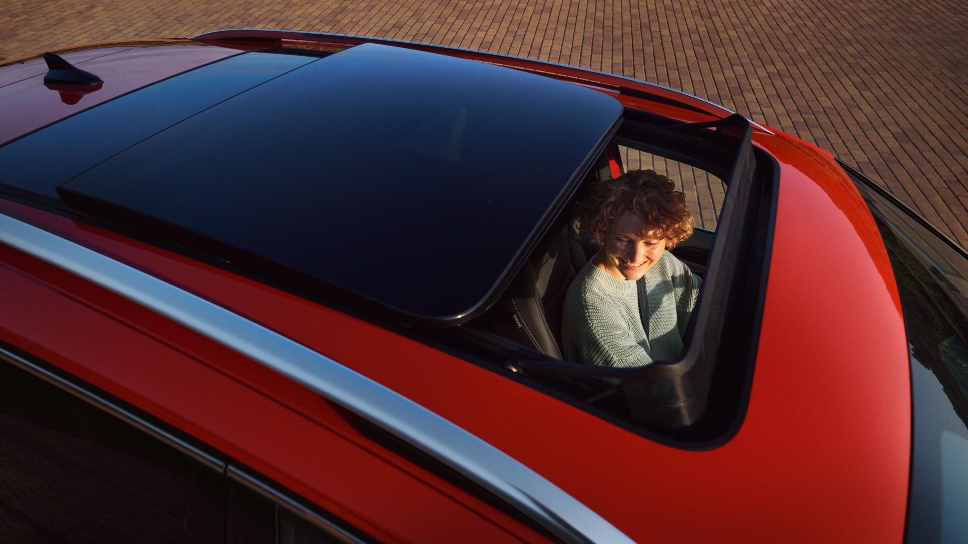 Topshot durch das halb geöffnete optionale Panorama-Ausstell-/ Schiebedach eines roten VW Tiguan. Auf dem Fahrersitz ist eine junge Frau zu sehen.