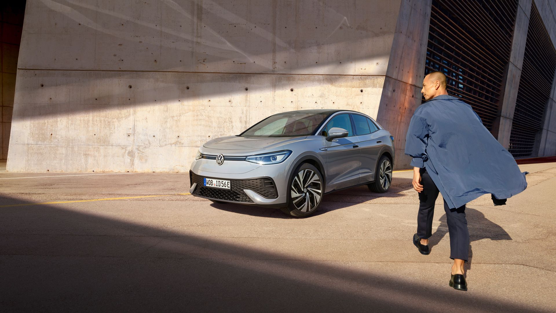 Grauer VW ID.5 parkt vor heller Hauswand, Ansicht schräg rechts von vorn, Mann geht zügig auf das E-Auto zu.