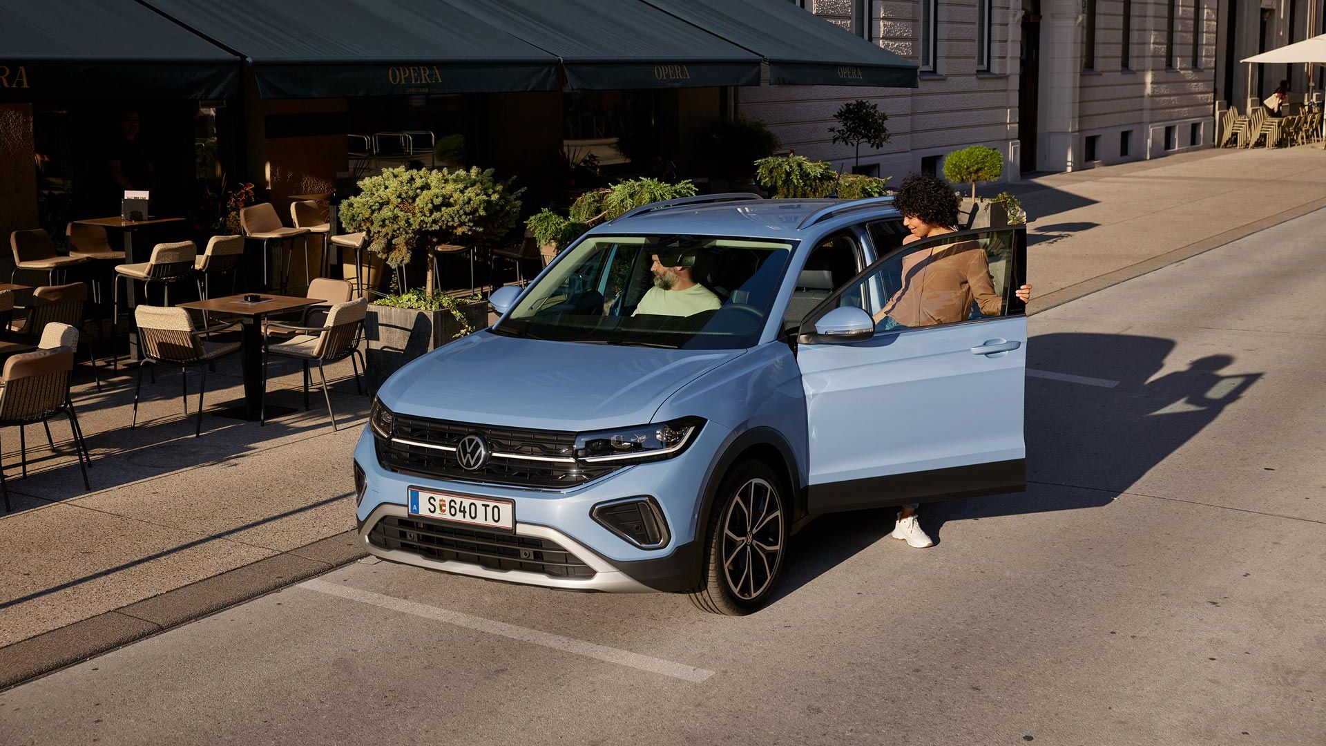 Eine Frau steigt auf der Fahrerseite in den blauen VW T-Cross, der vor einem urbanen Straßencafé parkt.