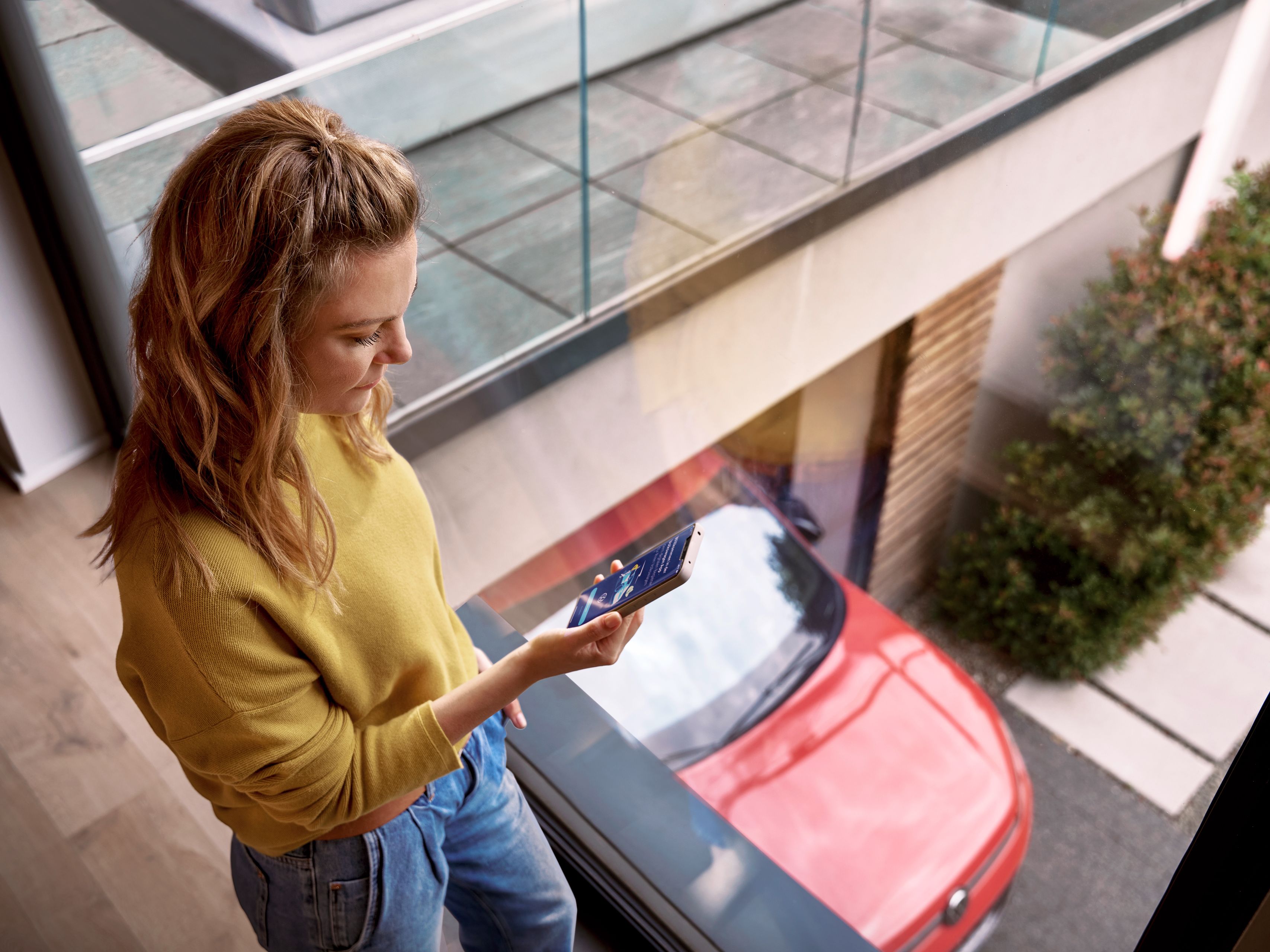 Žena gleda u mobilni telefon. U pozadini crveni Volkswagen. U desnom gornjem uglu logotipi App Store i Google Play Store prodavnice.