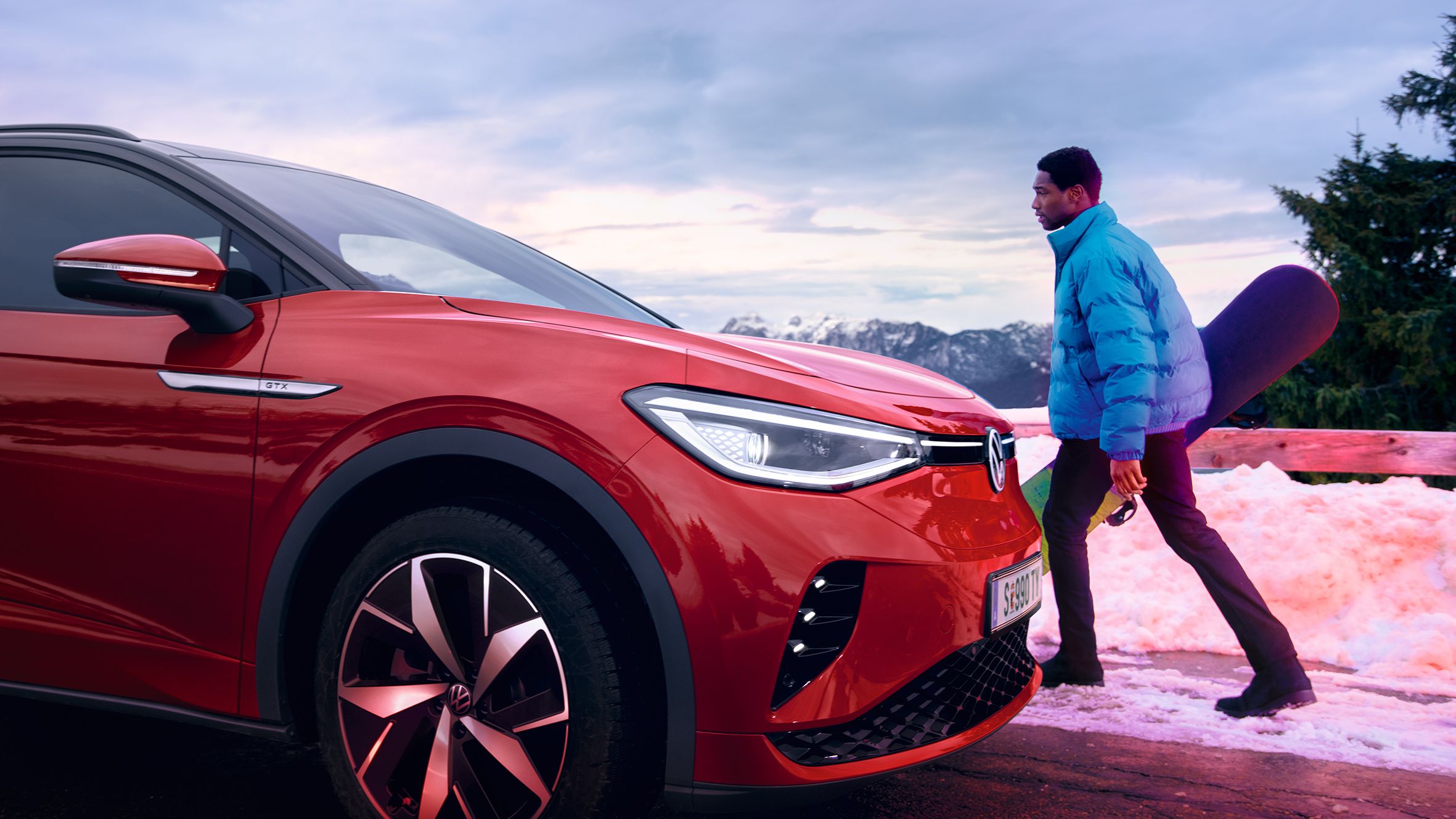 VW ID.4 GTX in rot von der Seite im Schnee auf Straße. Mann mit Winterjacke und Snowboard in der Hand geht auf das Fahrzeug zu.