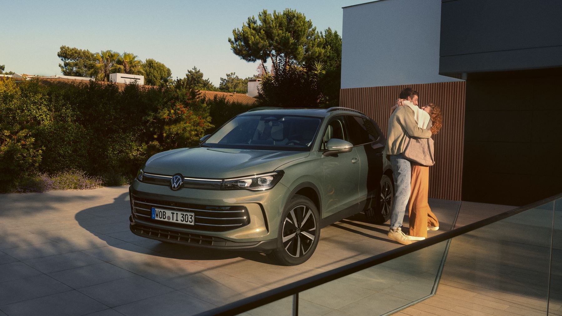 Grüner VW Tiguan parkt in Frontansicht auf der Einfahrt eines modernen Wohnhauses. Ein junges Paar steht daneben und umarmt sich.