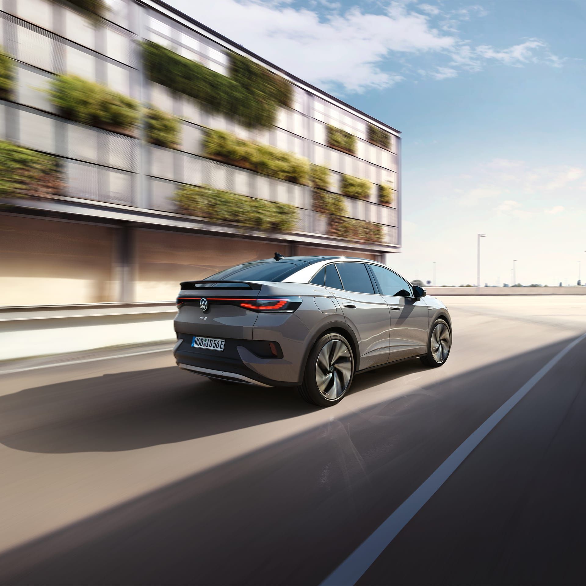 Fahraufnahme eines grauen VW ID.5 von schräg hinten, links ein helles, teilweise grün bewachsenes Gebäude