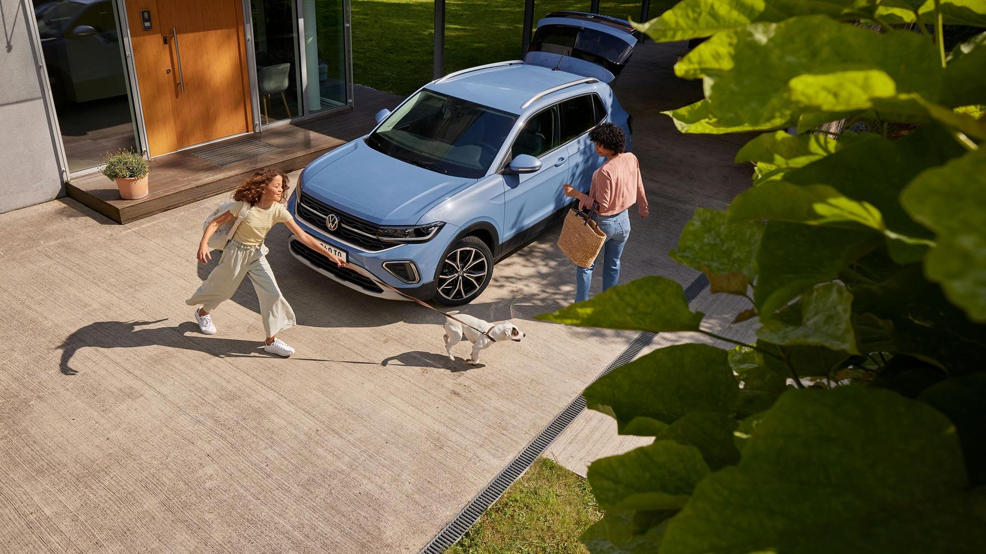 Eine Frau mit Korb und ein Mädchen mit Hund vor einem blauen VW T-Cross mit geöffnetem Kofferraum in einer Einfahrt.