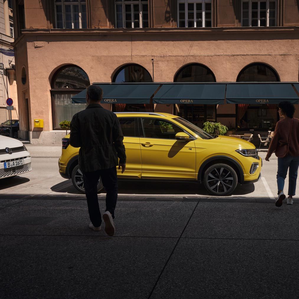 Mann und Frau nähern sich einem gelben VW T-Cross am Straßenrand.