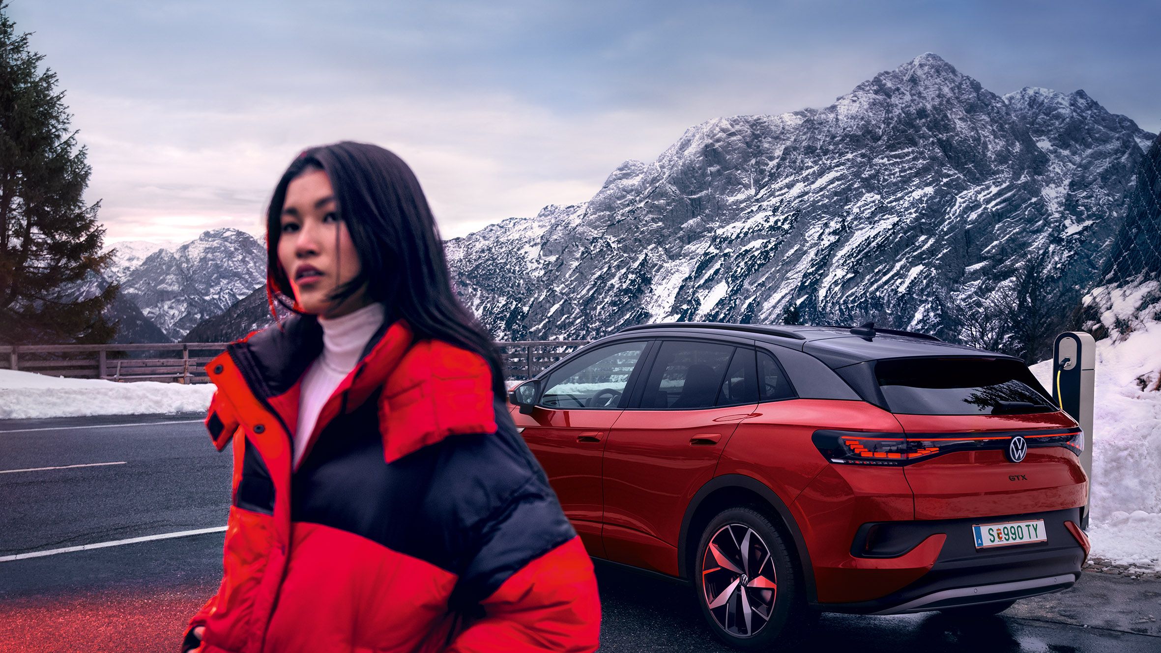 VW ID.4 GTX in rot auf Straße in Berglandschaft mit Schnee an Ladesäule. Frau in roter-schwarzer Jacke überquert die Straße.